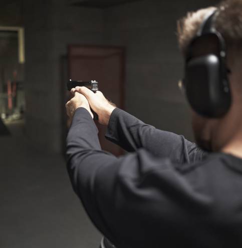 Caucasian young adult man wearing protective earmuffs practicing shooting at indoor firing range, holding handgun with both hands and aiming at paper targets in distance